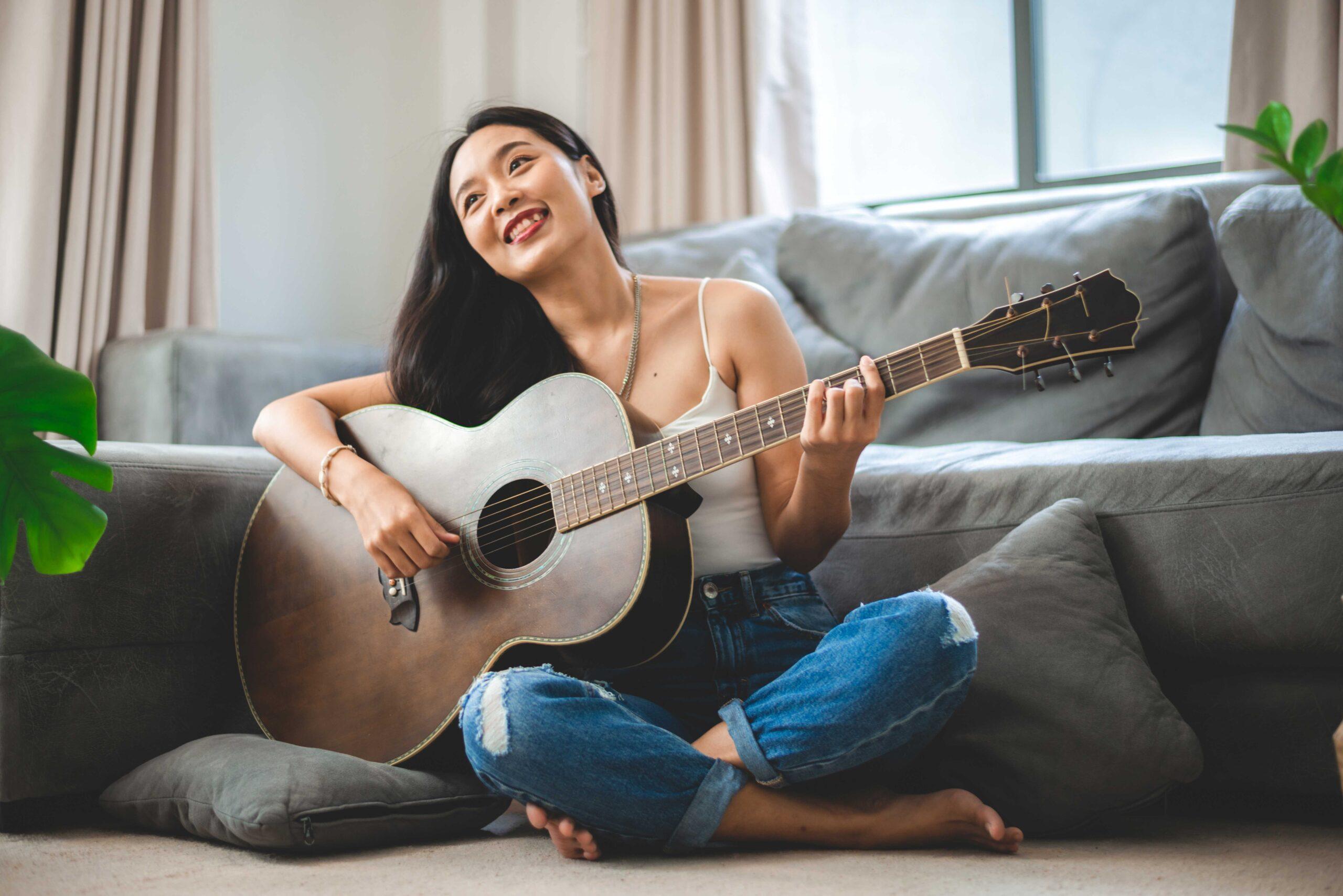 Asiatische Frau, die zu Hause Musik mit der Gitarre spielt, junge Gitarristin, Musikerin, Lebensstil mit akustischem Kunstinstrument, das sitzt, um zu spielen